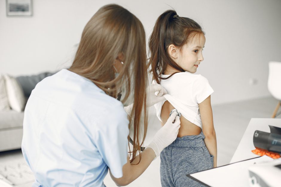 Is Your Family Due for a Well-Visit Before School Registration Closes? A female family doctor examines a child with a stethoscope in a medical exam room during a well-visit checkup.